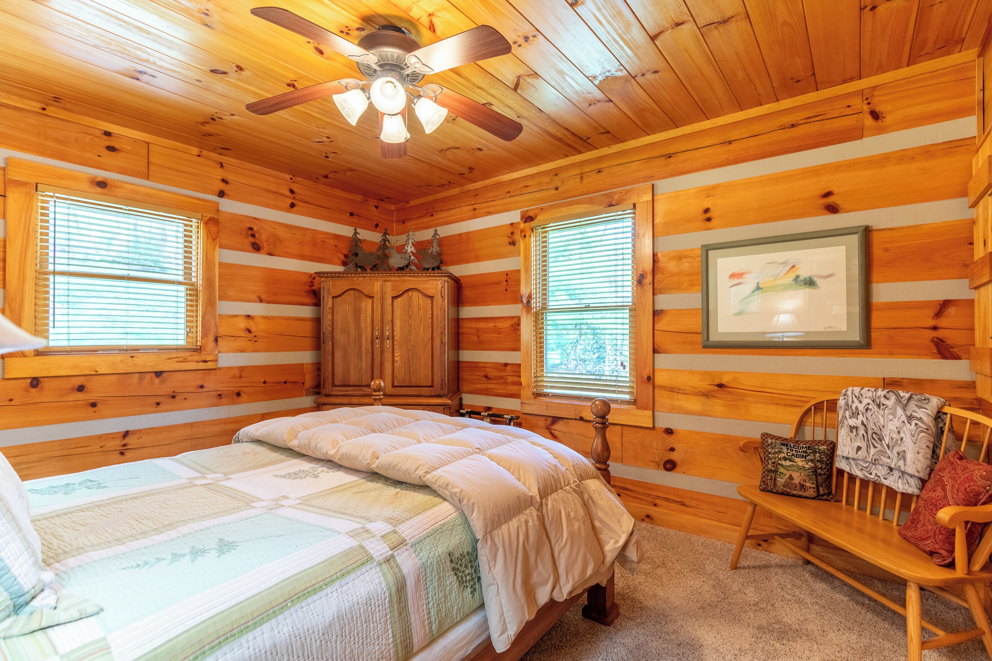Une chambre dans un chalet en rondins, avec un lit, un banc en bois, un ventilateur de plafond et un tableau encadré au mur.