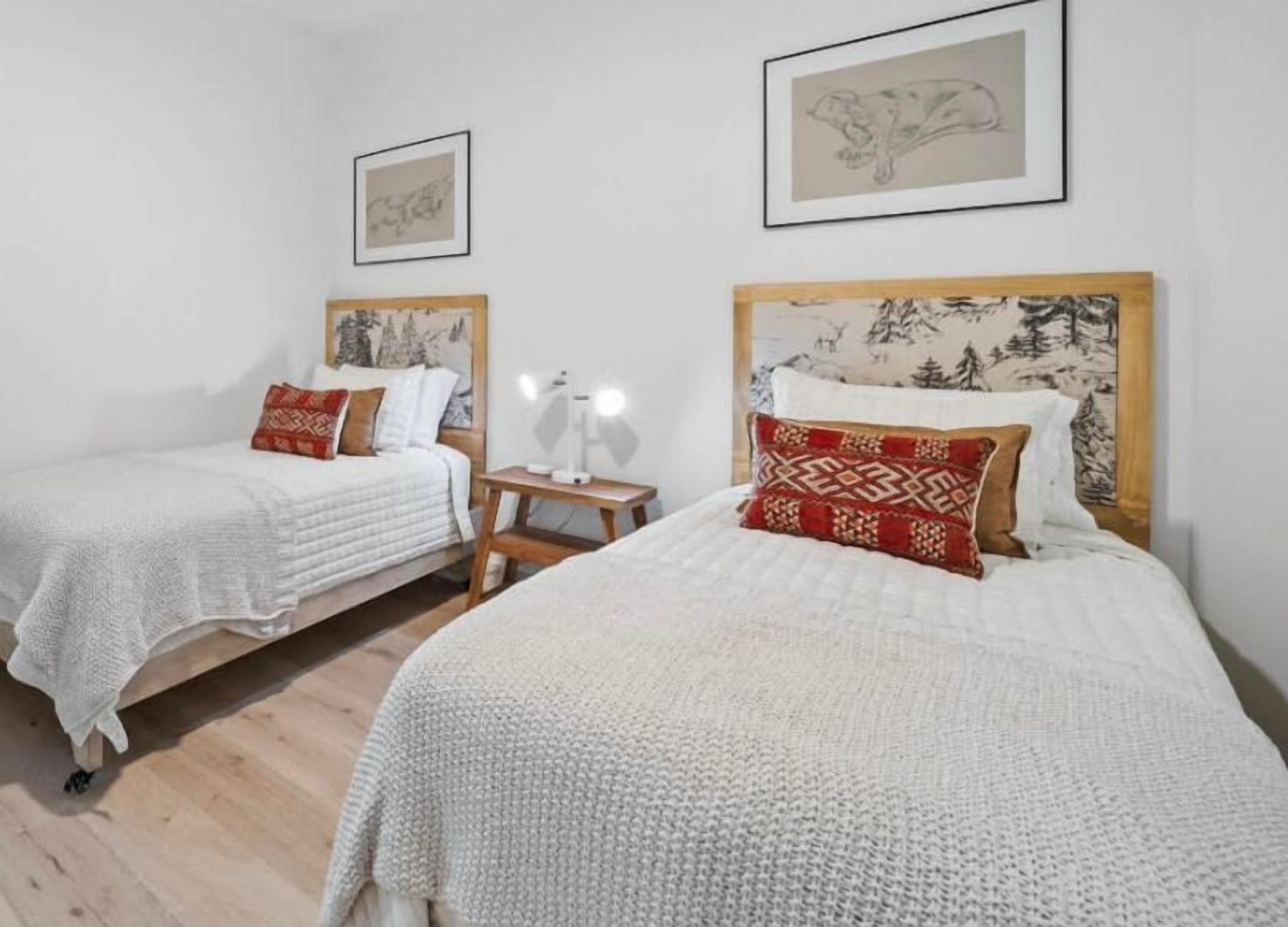 Deux lits simples avec du linge de lit blanc et des oreillers à motifs, une petite table de chevet avec une lampe, et des œuvres d’art encadrées accrochées aux murs.