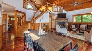 A spacious living area with a large wooden dining table, comfortable seating, a stone fireplace, and a staircase leading to an upper level. - Shady Pines (Banner Elk)