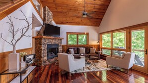A spacious living room with a stone fireplace, leather sofas, a flat-screen TV, and large windows offering a view of the outdoors. - Shady Pines (Banner Elk)