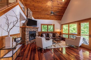 A spacious living room with a stone fireplace, leather sofas, a flat-screen TV, and large windows offering a view of the outdoors. - Shady Pines (Banner Elk)