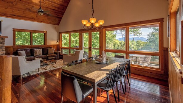 A spacious dining area with a wooden table, chairs, and a chandelier. Large windows offer views of trees and mountains. - Shady Pines (Banner Elk)