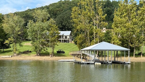 A lakeside gazebo with a dock extending into the water, surrounded by trees and a hill with a house in the background. - Sweet Water Estate (Butler)