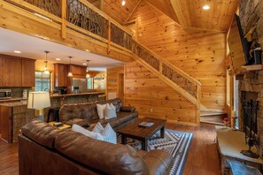 Ein geräumiges Blockhaus-Interieur mit Wohnbereich, der ein Ledersofa, einen Couchtisch und einen Kamin umfasst. Eine Holztreppe mit Geländer führt auf eine erhöhte Ebene.