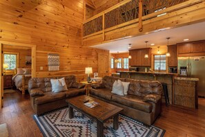 Ein geräumiges Holzhaus im Inneren mit einem Wohnbereich, der zwei Ledersofas, einen Couchtisch und einen gemusterten Teppich beherbergt. Im Hintergrund ist eine Küche mit Holzausstattung und Edelstahlgeräten zu sehen.