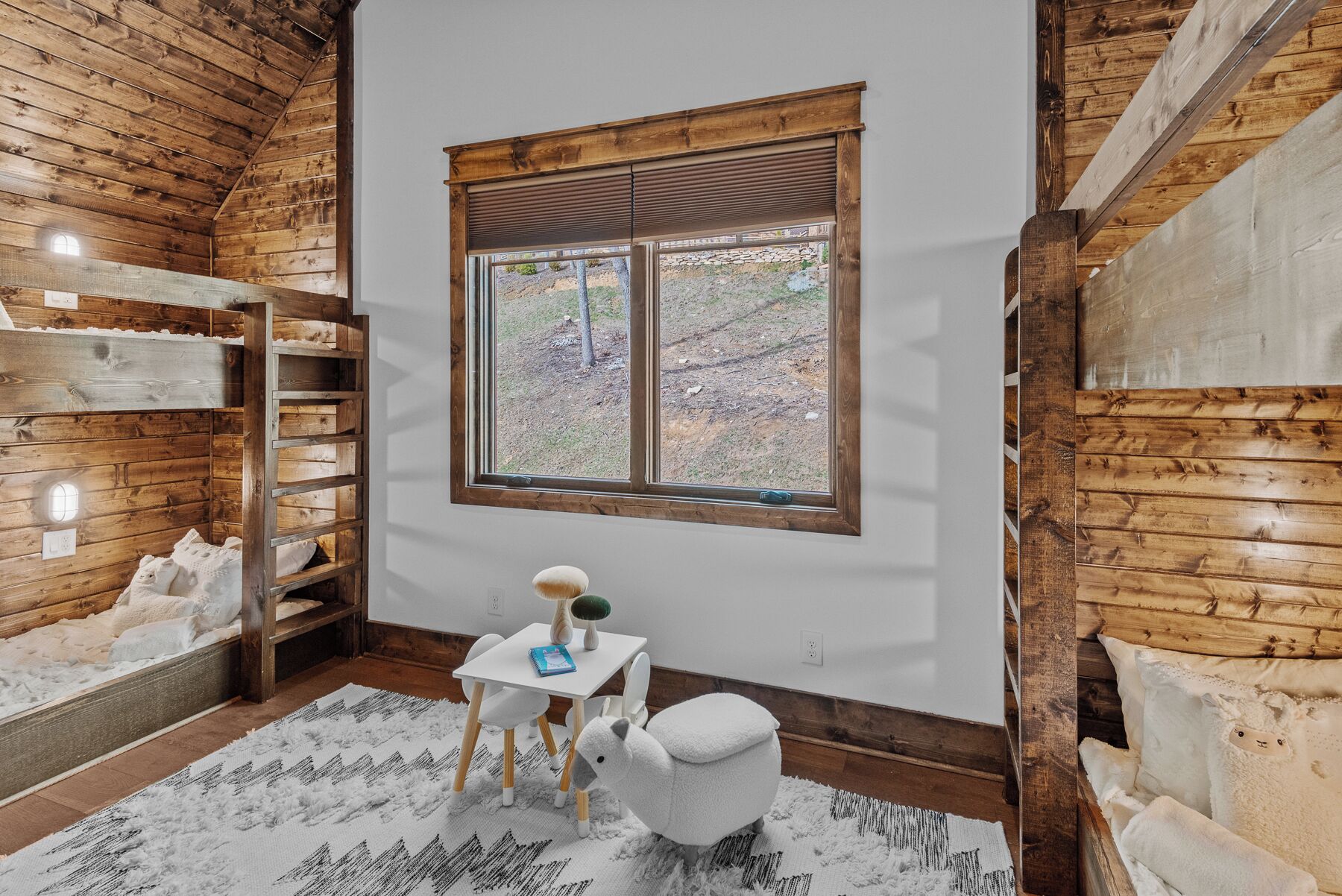 Ein gemütliches Zimmer mit Holzwänden, einem Fenster mit Jalousien, einem weißen Tisch mit Lampe, einem weißen Stuhl und einem gemusterten Teppich.