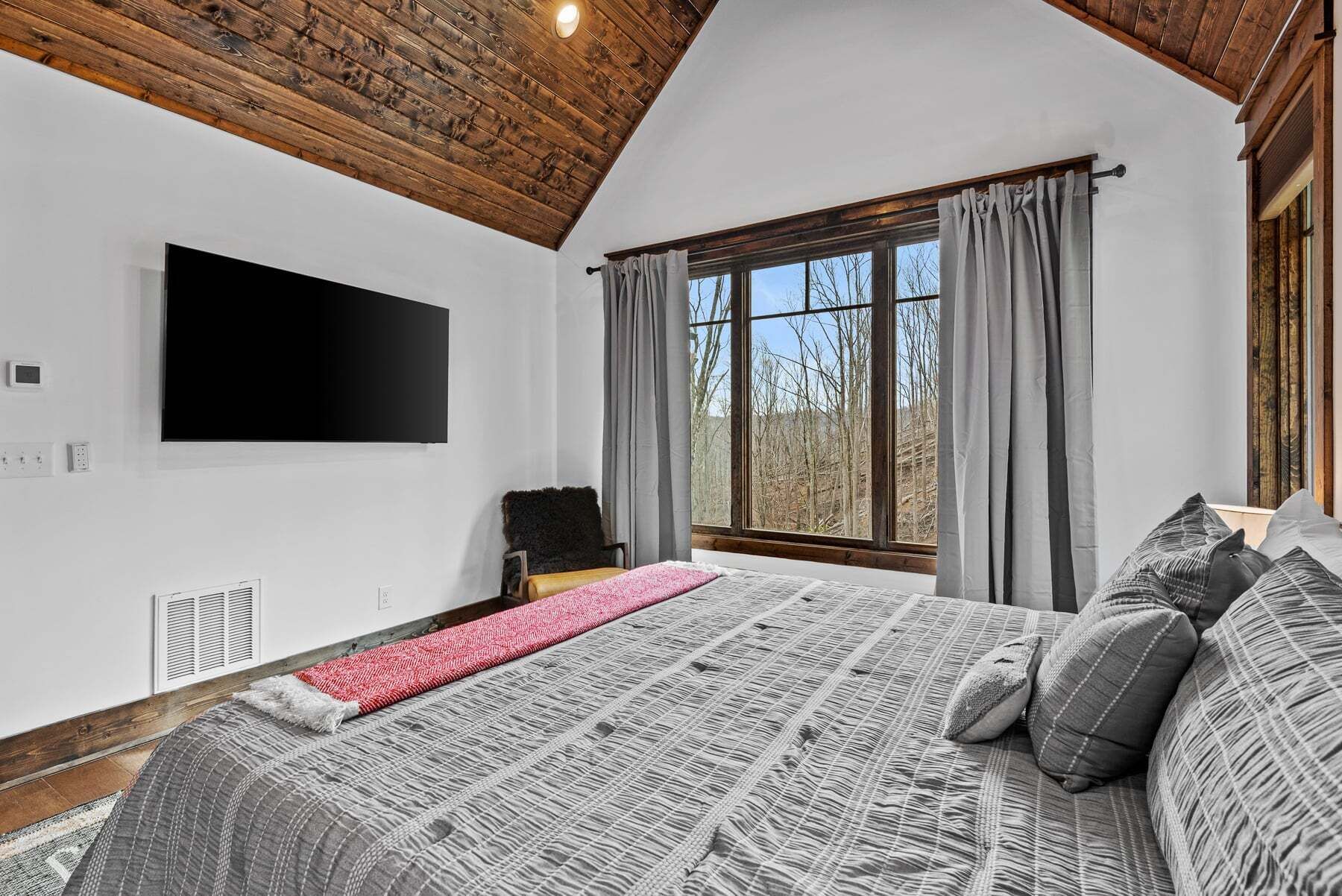 A bedroom with a large bed, a flat-screen TV, a window with curtains, and a wooden ceiling.