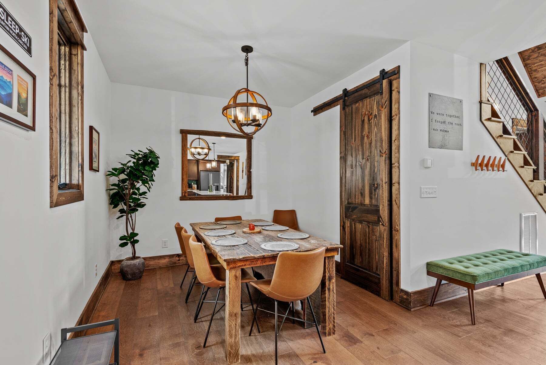 A dining area with a wooden table, chairs, a hanging light fixture, a large mirror, and a staircase with a wooden door.