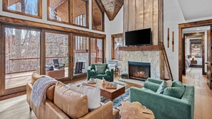 A spacious living room with large windows, a stone fireplace, and a television mounted on the wall. - Llama Creek Cabin at Eagles Nest (Banner Elk)
