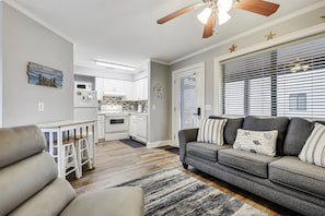 A modern living room with a gray sofa, a wooden ceiling fan, and a kitchen in the background. - Holiday Hideaway - Sand Castle Villas #30 (Panama City Beach)