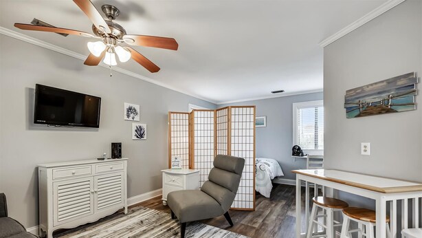 A modern living room with a ceiling fan, a TV, a white cabinet, a grey armchair, a wooden partition, and a small table with stools. - Holiday Hideaway - Sand Castle Villas #30 (Panama City Beach)