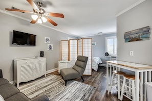 A modern living room with a ceiling fan, a TV, a white cabinet, a grey armchair, a wooden partition, and a small table with stools. - Holiday Hideaway - Sand Castle Villas #30 (Panama City Beach)