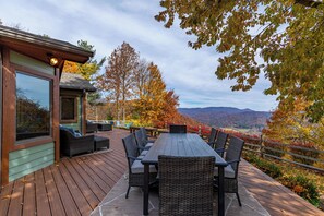 Eine Holzterrasse mit Gartenmöbeln, einem großen Fenster und Blick auf eine Berglandschaft.