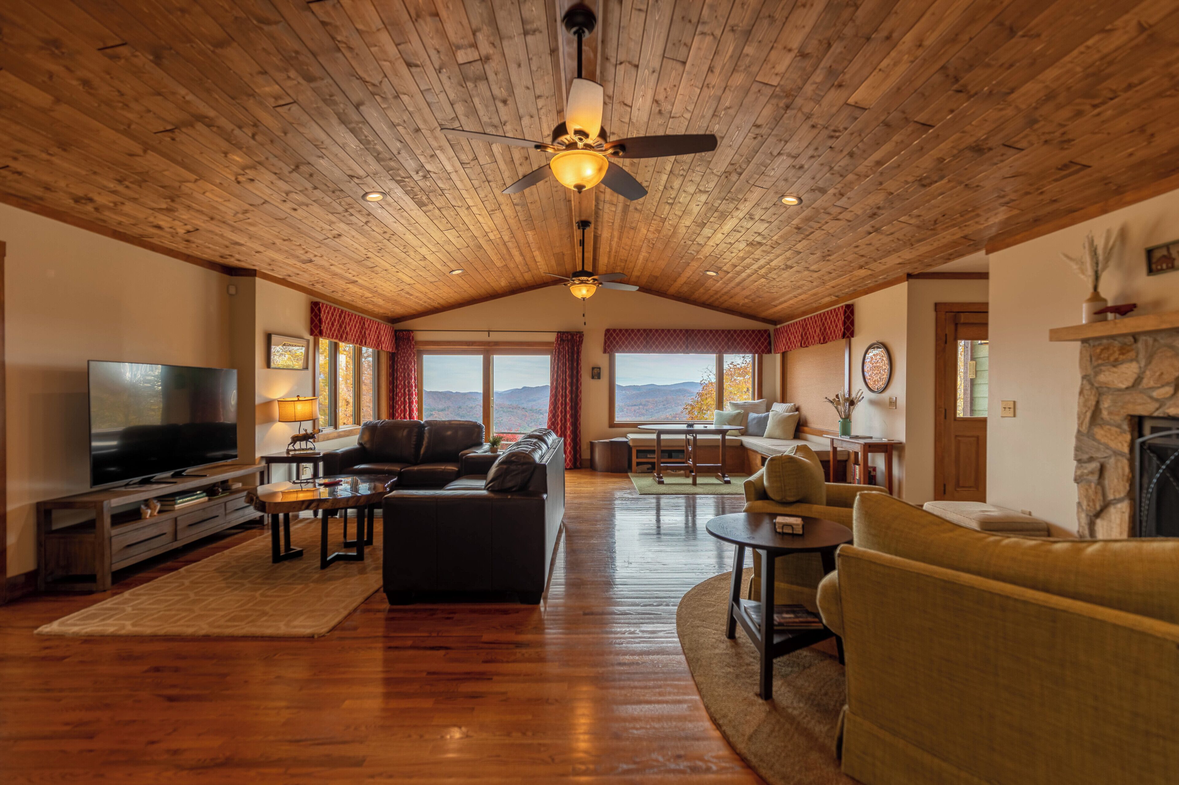 A spacious living room with a wooden ceiling, a large flat-screen TV, leather sofas, and a stone fireplace.