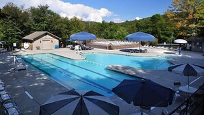 Piscina com espreguiçadeiras e guarda-sóis, rodeada por árvores e um pequeno edifício.