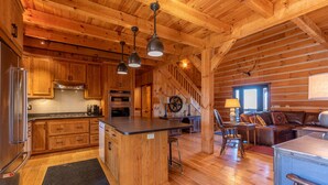 Ein Holzblockhaus-Interieur mit Holzdielenböden, einer Küche mit Holzschränken, einer zentralen Kochinsel, einer Treppe, einem Wohnbereich mit Sofa und Lampe sowie einem Rehgeweih an der Wand.