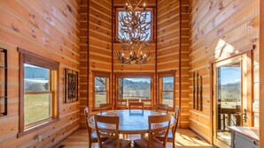 Ein Holzhütten-Interieur mit einem runden Esstisch, Holzstühlen, einer Chandelüster und großen Fenstern, die Ausblicke ins Freie bieten.