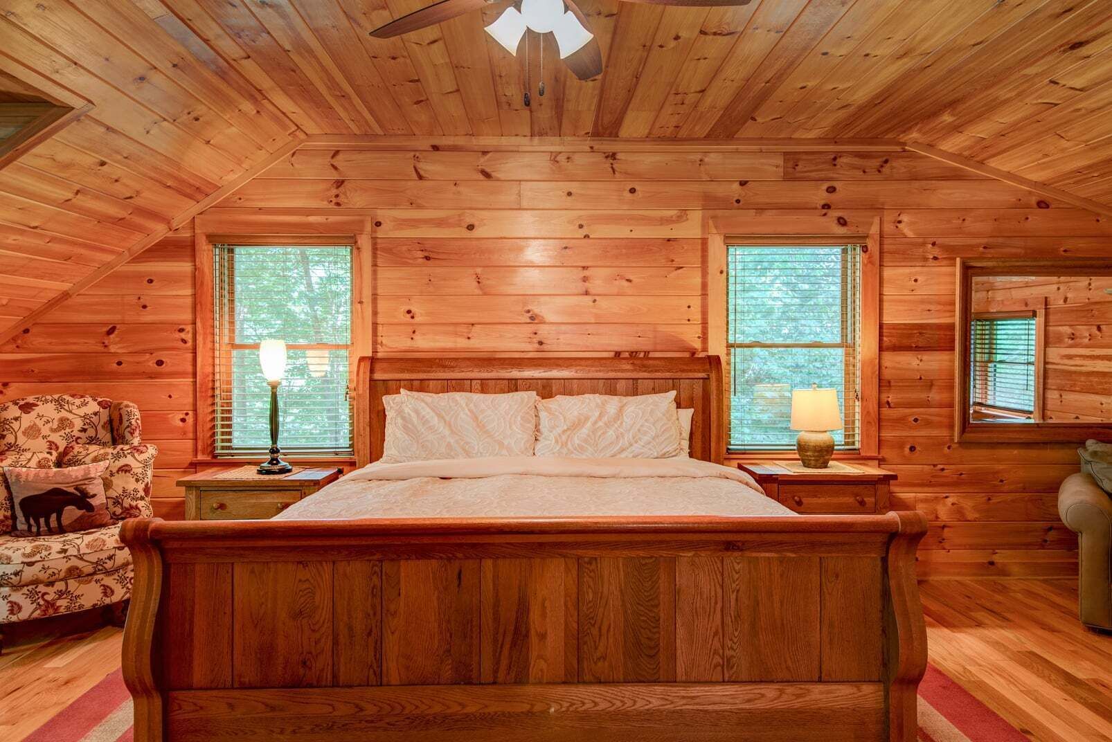 Un dormitorio tipo cabaña de madera con cama, mesitas de noche, una silla y un ventilador de techo.
