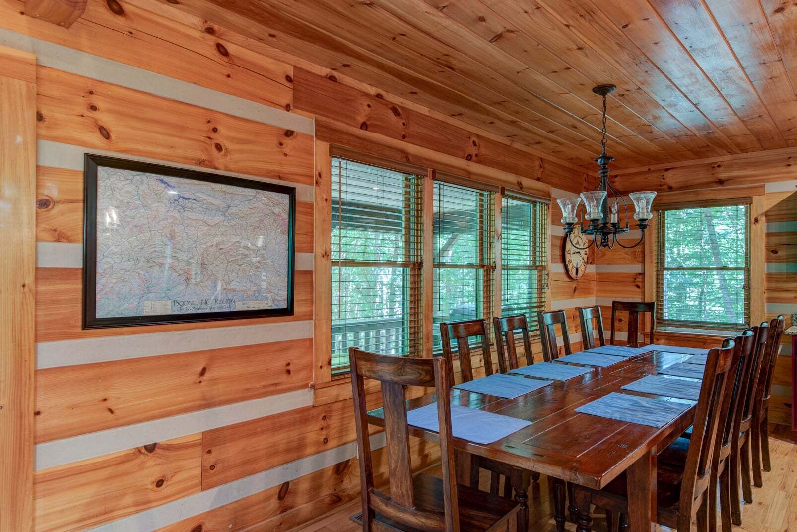 A wooden cabin interior with a dining table, chairs, a map on the wall, and large windows.