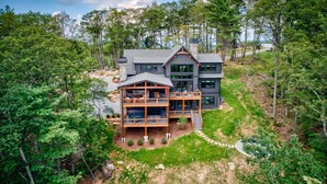 A large, multi-story house with a deck, surrounded by dense forest and a clear sky. - One More Day at Eagles Nest (Elk Park)