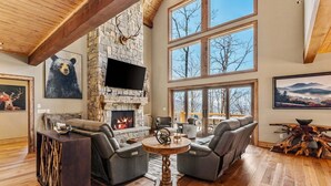 A spacious living room with a stone fireplace, leather sofas, a flat-screen TV, and large windows offering a view of trees and mountains. - Rock Haven at Eagles Nest (Banner Elk)