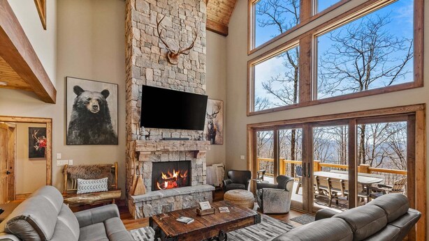 A stone fireplace with a mounted deer head, a flat-screen TV, leather furniture, and large windows overlooking a deck. - Rock Haven at Eagles Nest (Banner Elk)