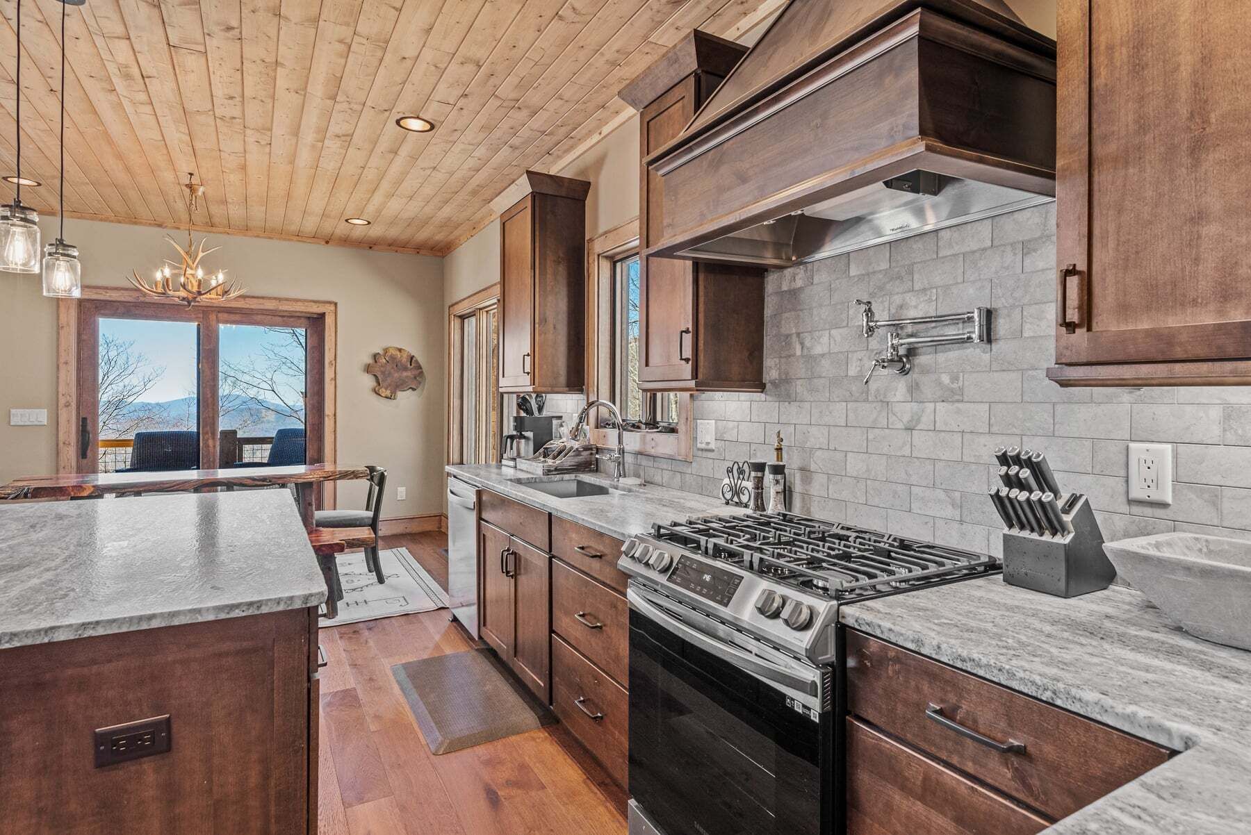 A kitchen with wooden cabinets, a gas stove, a dishwasher, and a large window overlooking a mountainous landscape.