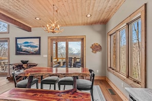 A dining area with a wooden table, chairs, a chandelier, a painting, and a large window. - Rock Haven at Eagles Nest (Banner Elk)