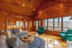 A spacious living room with a large sectional sofa, two green armchairs, a wooden coffee table, and a round rug. The room features wooden walls and ceiling, large windows offering a view of the outdoors.