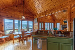 A wooden cabin interior with a dining area, kitchen, and large windows offering views of the outdoors.