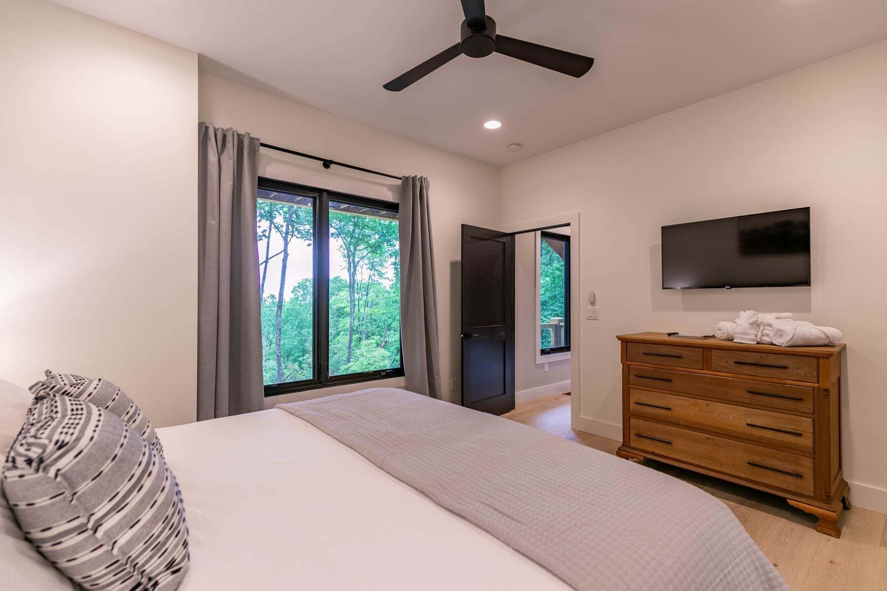 Un dormitorio con cama, cómoda, ventilador de techo, televisor y ventana con vista a árboles.