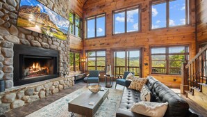 A log cabin interior with a stone fireplace, a leather sofa, and large windows offering mountain views. - Cranberry View Lodge (Newland)