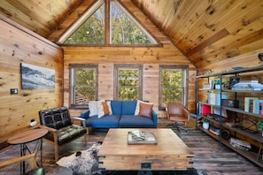 A cozy cabin interior with a wooden ceiling, a blue sofa, a leather chair, a coffee table, and a bookshelf.