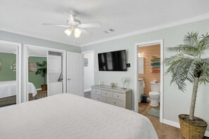 Quarto com cama, ventoinha no teto, televisão, cômoda, planta em vaso e banheiro visível através de uma porta aberta.