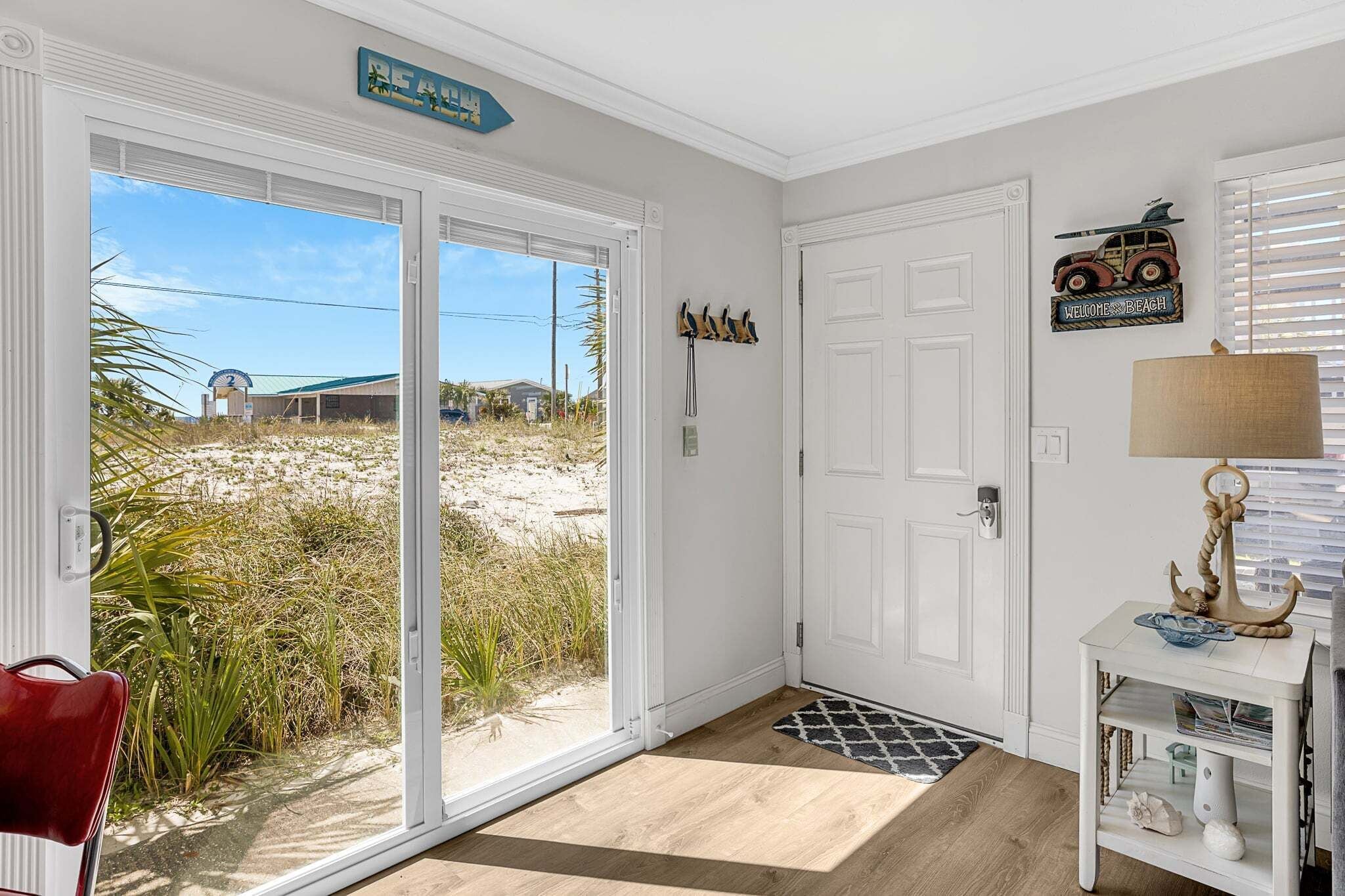 A beachside room with a sliding glass door, a door with a 'Welcome Beach' sign, a lamp, and a small table with decorative items.