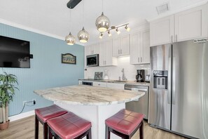 A kitchen with white cabinets, a stainless steel refrigerator, a marble island with red stools, pendant lights, and a flat-screen TV.