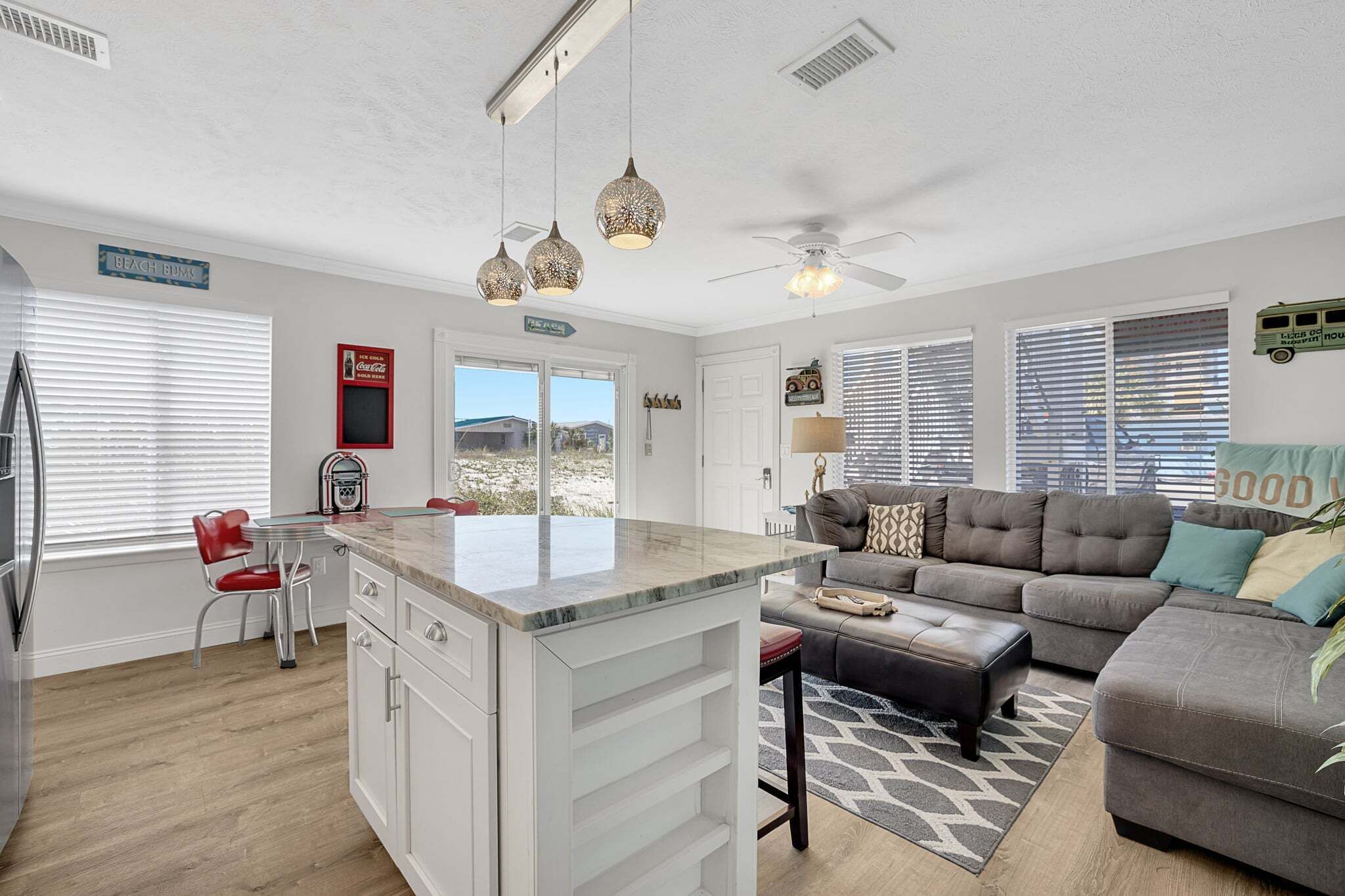 A modern kitchen with a central island, a dining area with a red chair, a sofa with a patterned rug, and a ceiling fan.