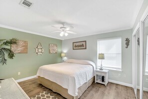 Quarto com cama, ventoinha no teto, decoração nas paredes, janela com persianas e mesa de cabeceira.