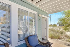 A covered porch with a wicker chair, a door, and a view of a sandy beach.