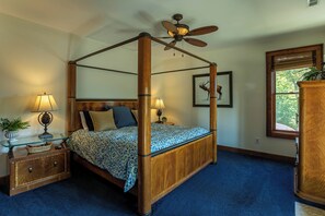 A four-poster bed with a ceiling fan, bedside lamps, a nightstand, and a window with a view of trees.