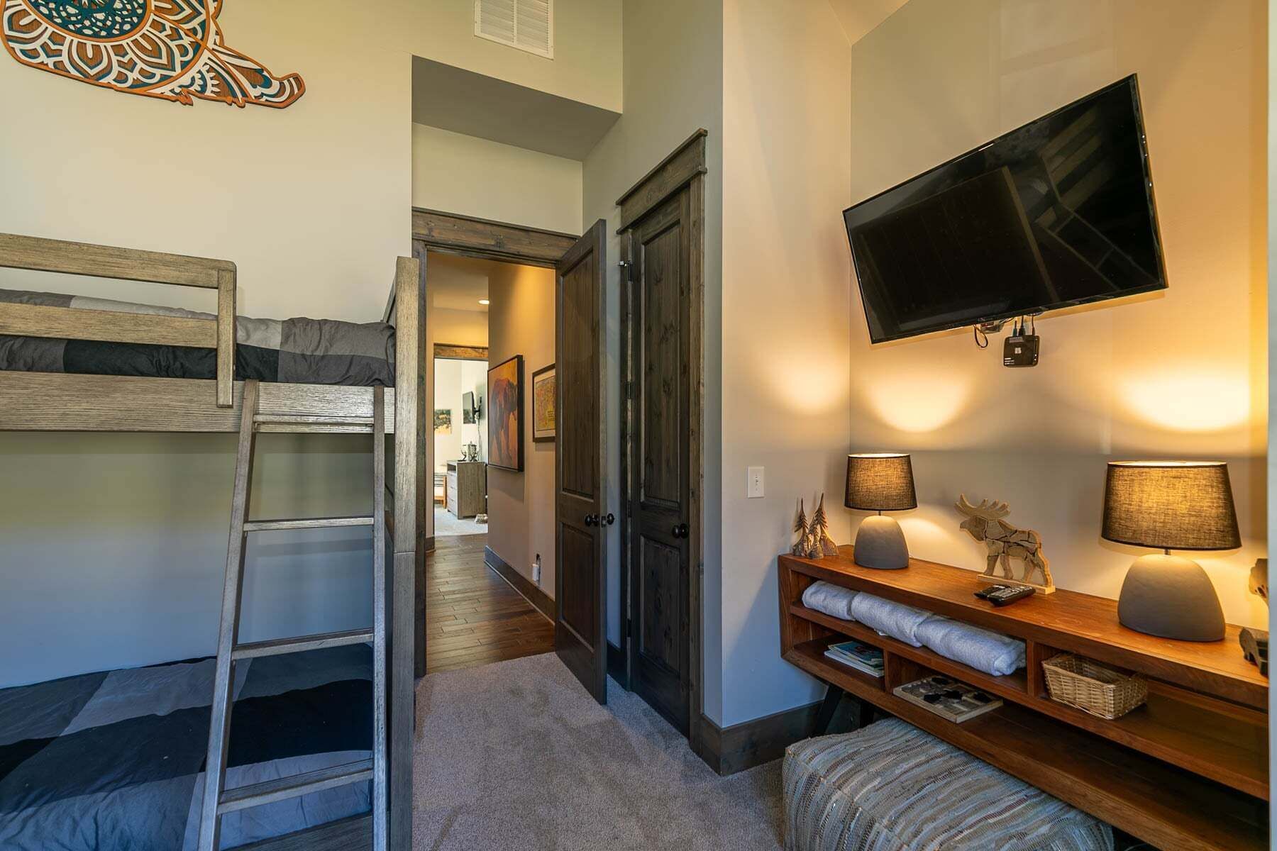 A bedroom with a bunk bed, a ladder, a TV mounted on the wall, and a bedside table with lamps.