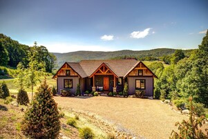 A two-story log cabin with a front porch, surrounded by a gravel driveway and a forested landscape.