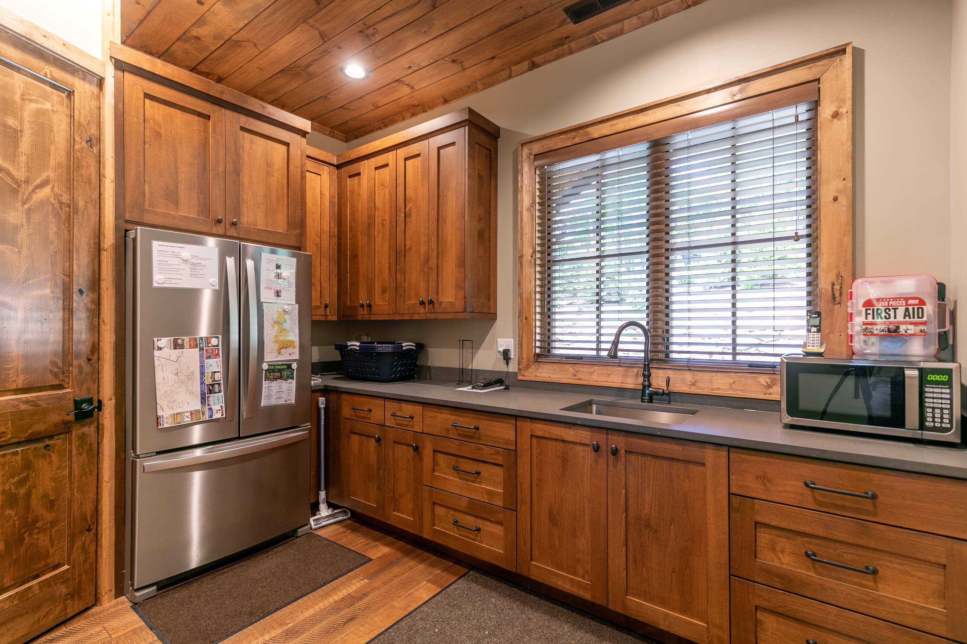 A kitchen with wooden cabinets, a stainless steel refrigerator, a microwave, and a window with blinds.