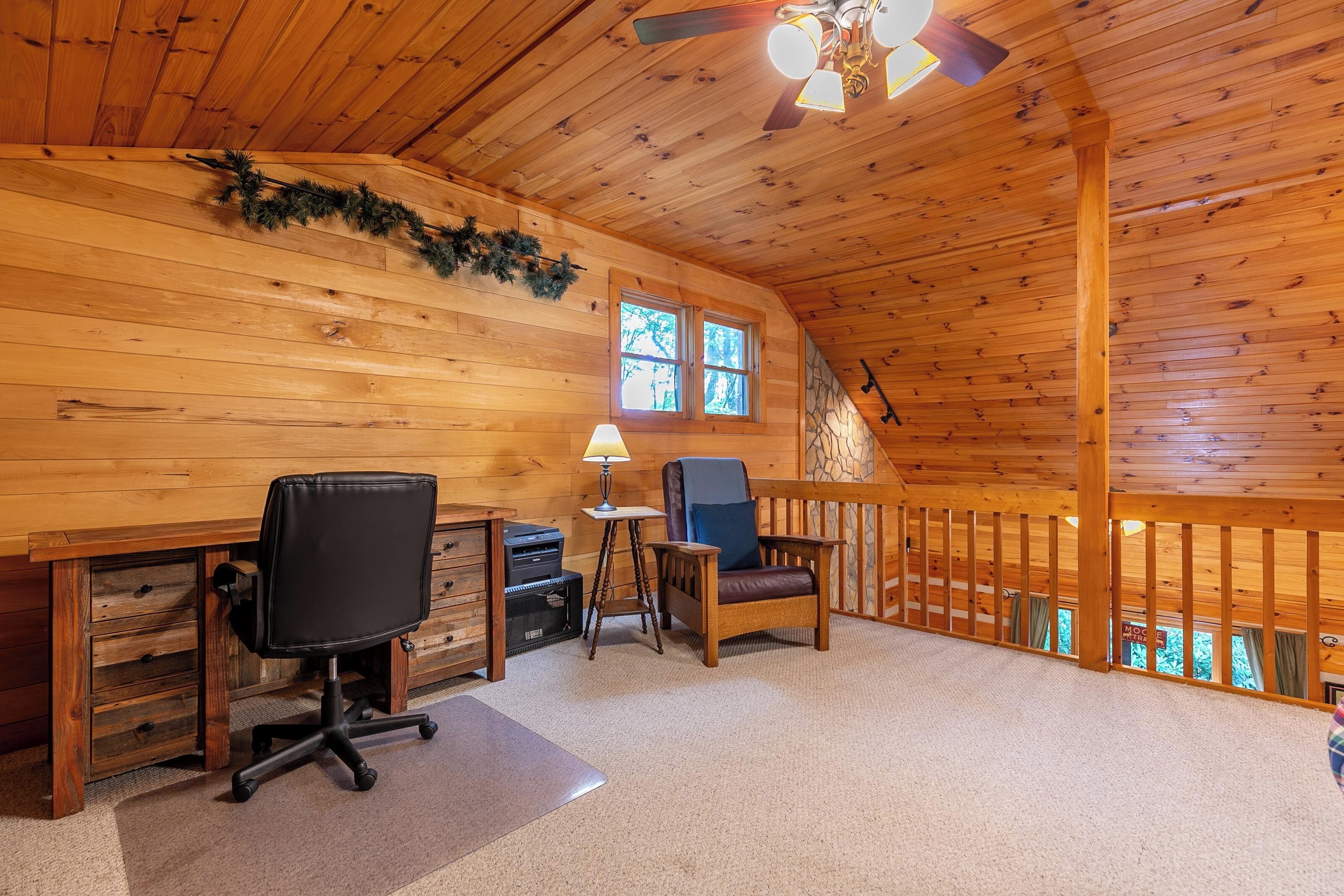 A cozy cabin interior with wooden walls, a desk with office chair, a small table with lamp, and a balcony with a railing.