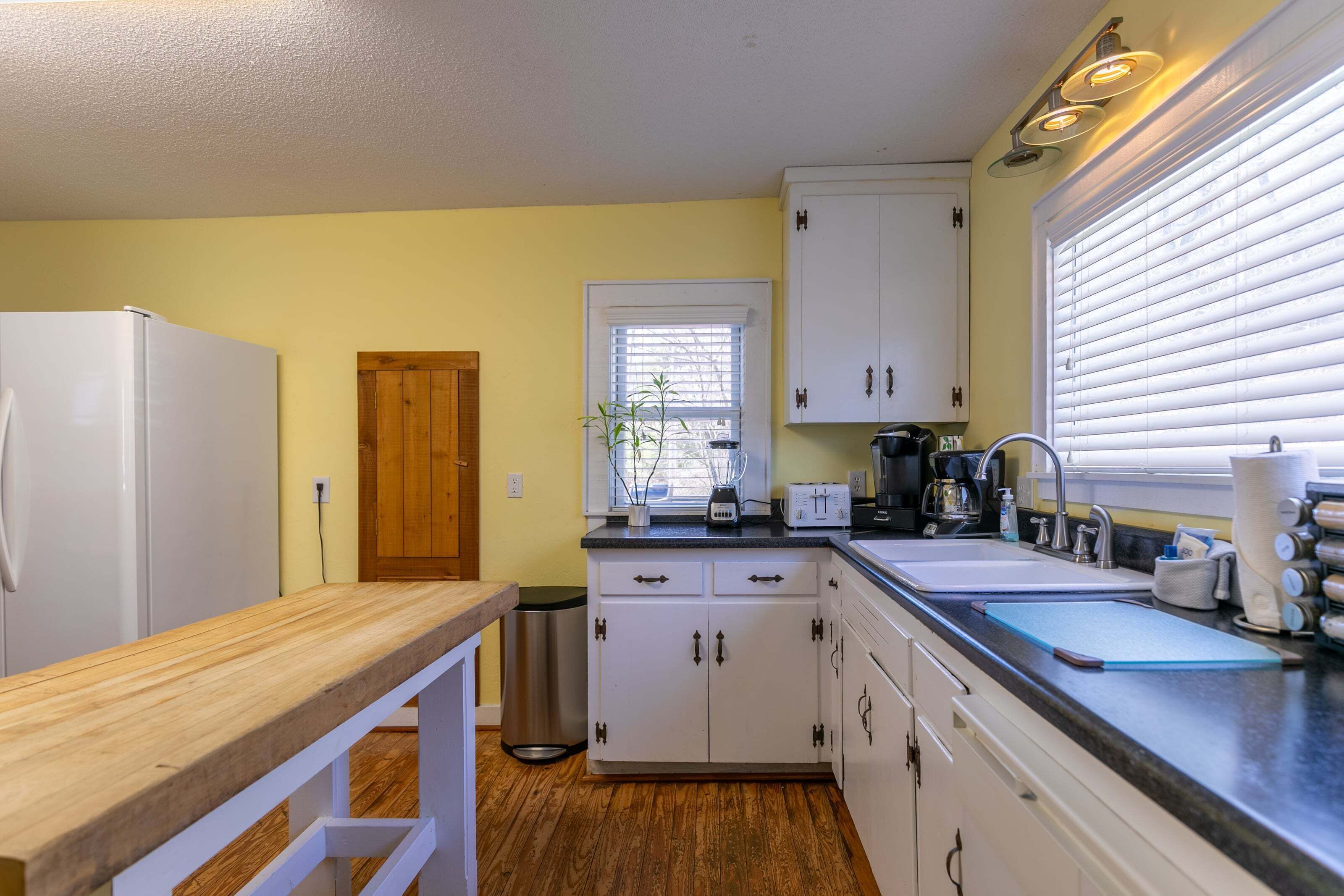 A compact kitchen with white cabinets, a wooden countertop, a window with blinds, and a refrigerator.