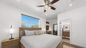 Un dormitorio con una cama grande, mesitas de noche, ventilador de techo, ventana y vistas al exterior.