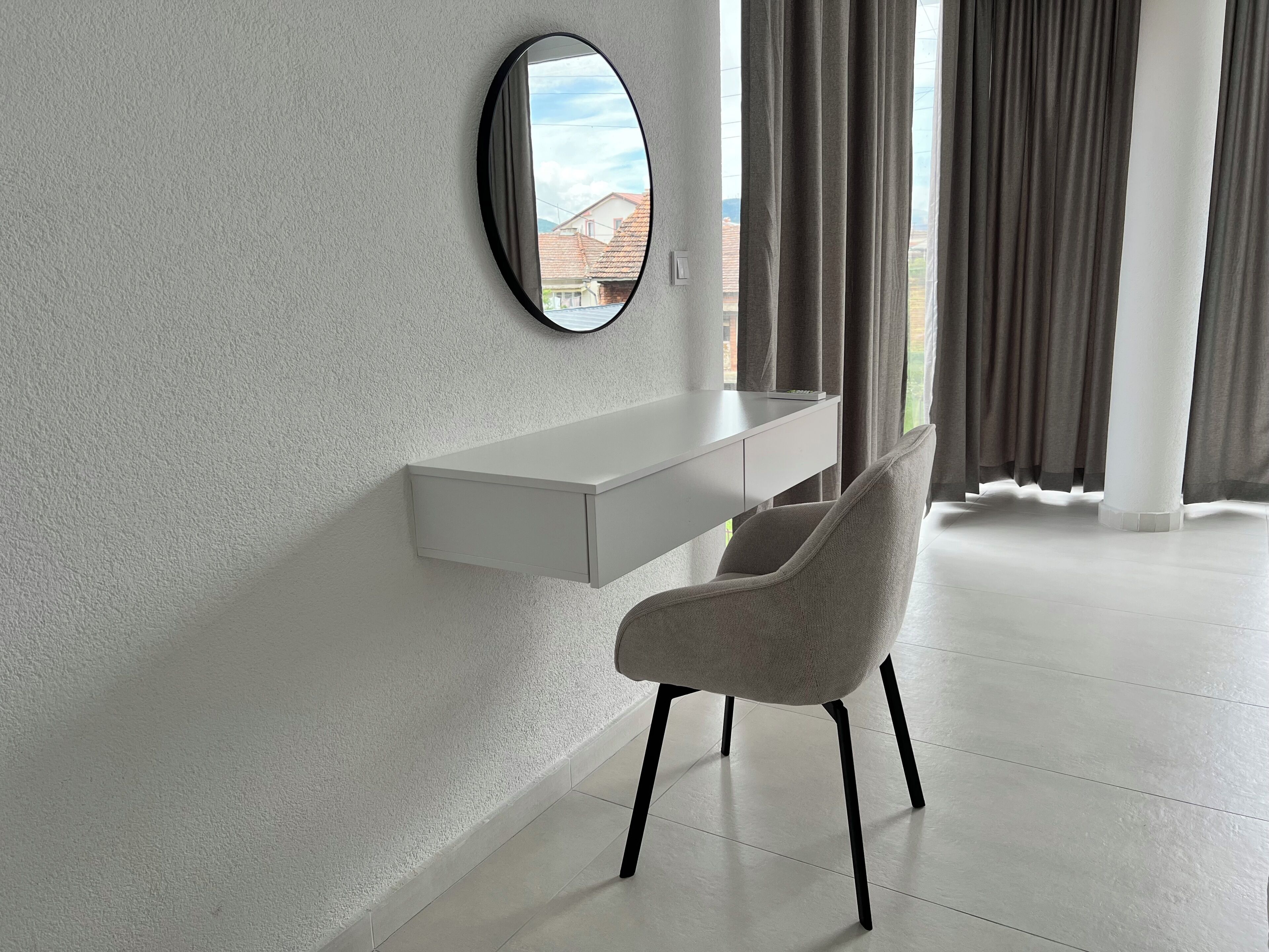 A modern room with a white desk, a grey chair, a round mirror, and a window with curtains.