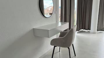 A modern room with a white desk, a grey chair, a round mirror, and a window with curtains.