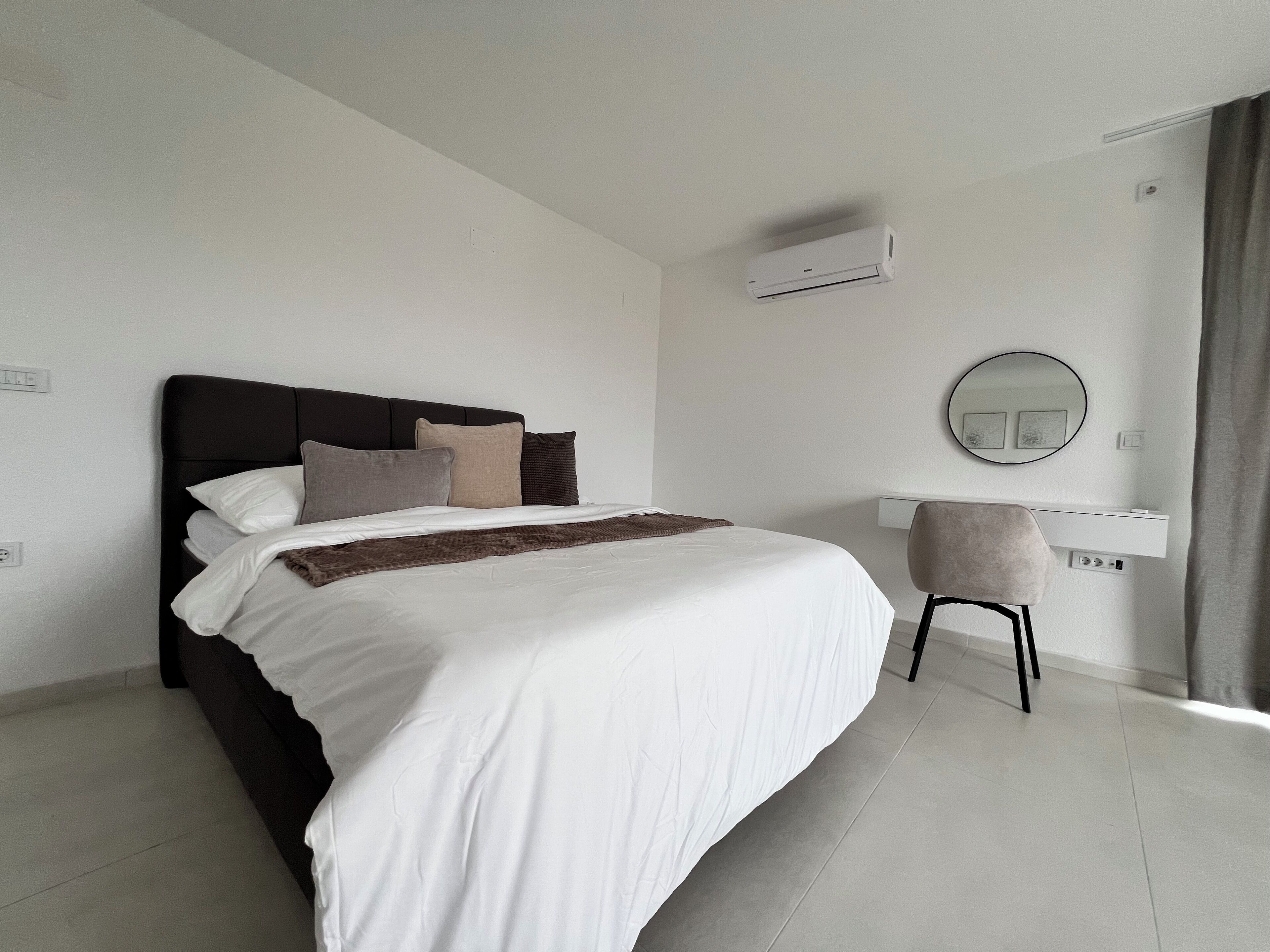 A modern hotel room with a large bed, a bedside table, a chair, and an air conditioning unit.