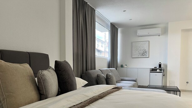 A modern hotel room with a large bed, a sofa, a desk, and a window with curtains.